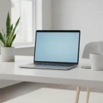 **Alt text:** Minimal home office workspace with an open laptop on a white desk, a green plant, and a coffee mug in a bright room.